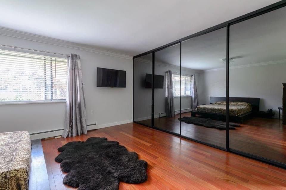 A bedroom with wood flooring, a large mirrored closet, a bed, a wall-mounted TV, two windows with gray curtains, and a black rug on the floor.